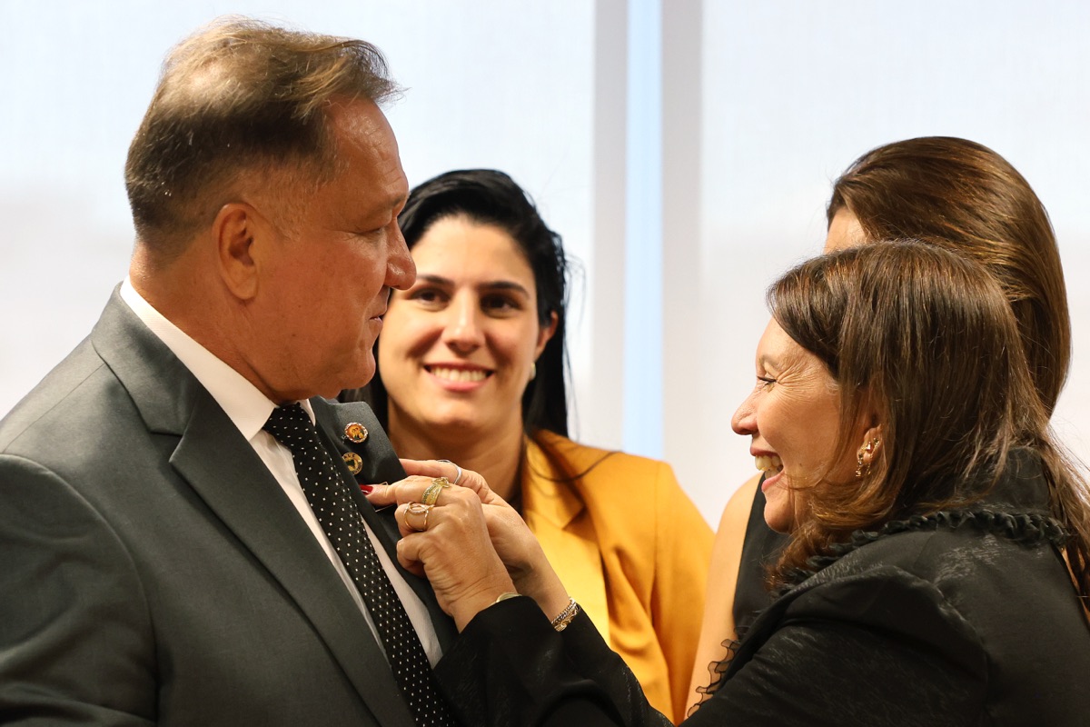 João Cardoso sendo cumprimentado por uma mulher em evento institucional, com outras pessoas ao fundo