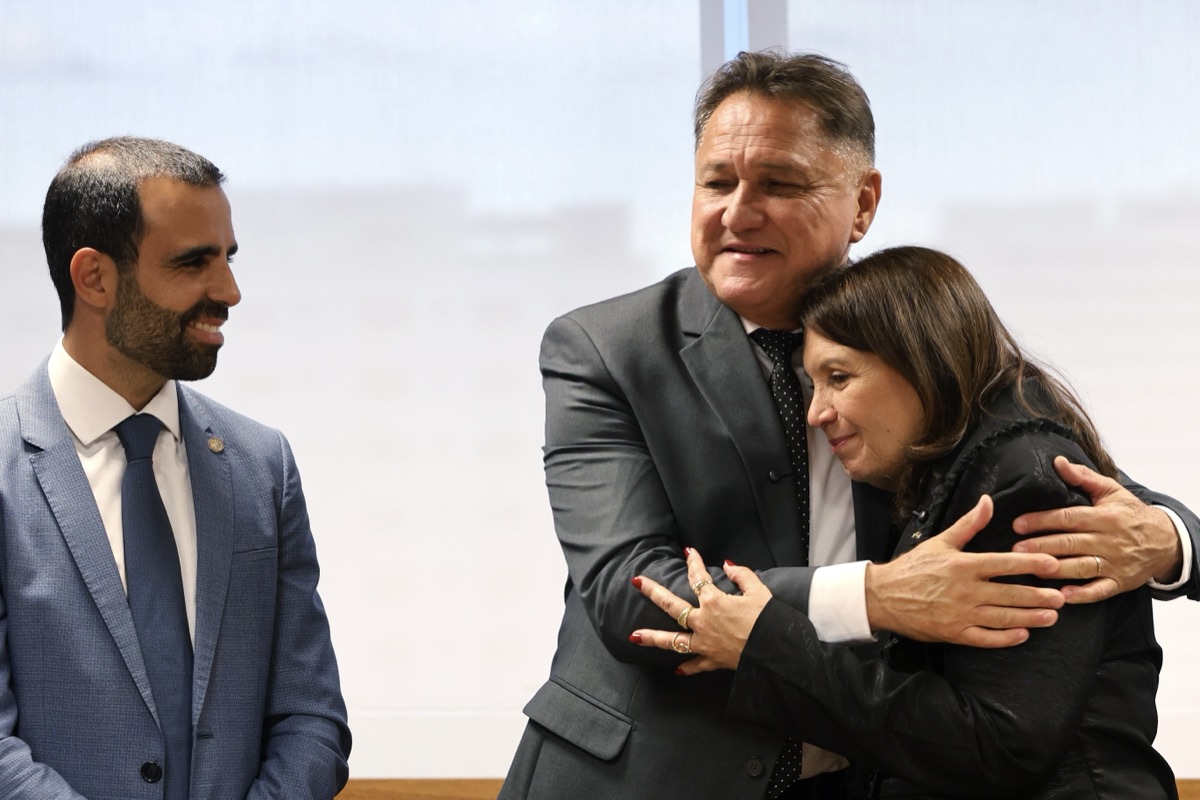 João Cardoso abraçando uma mulher, com outro homem de terno ao lado, em evento institucional