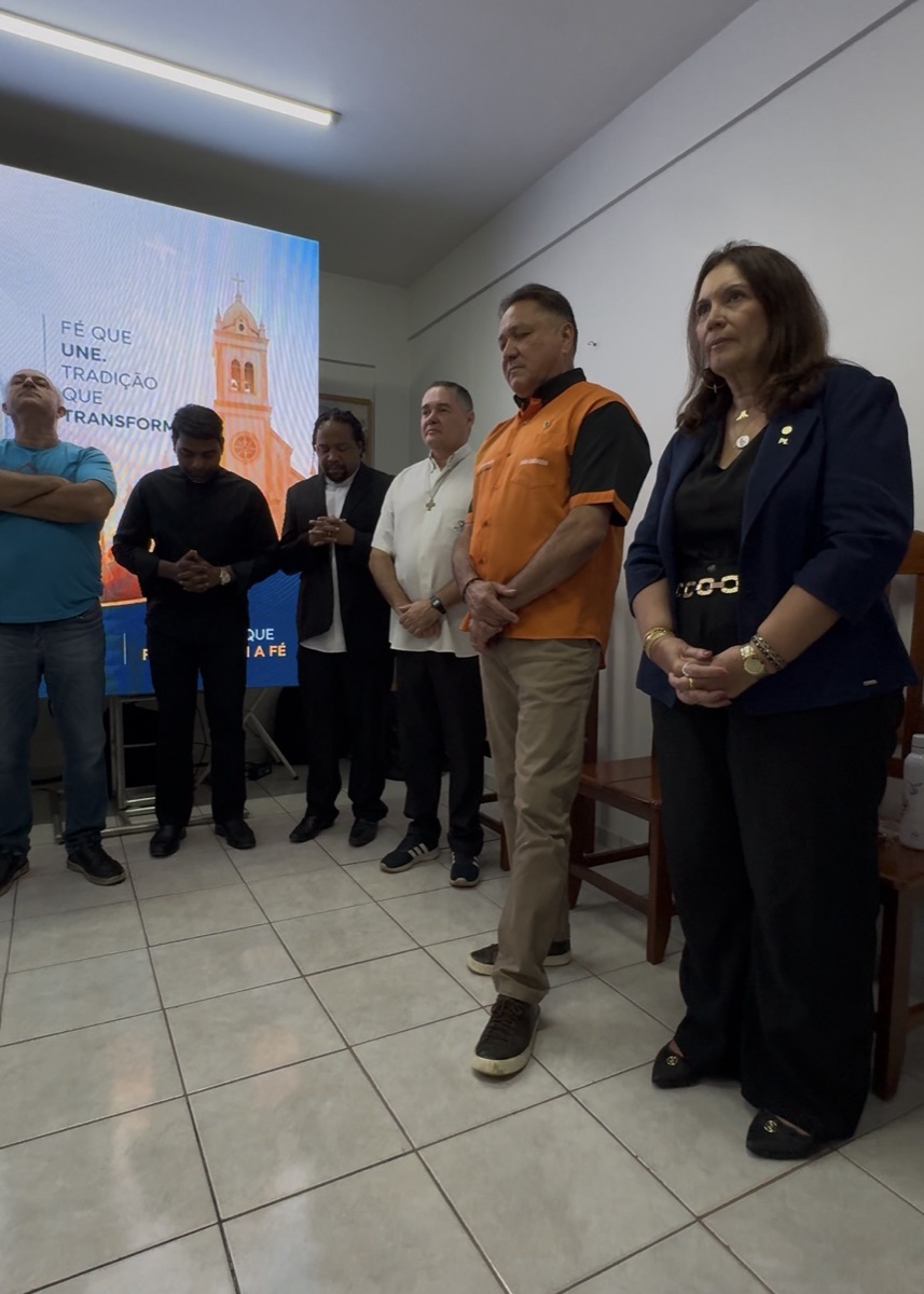 João Cardoso e Bia Kicis ao lado de padres e lideranças, com telão de fundo escrito "Fé que une, tradição que transforma"